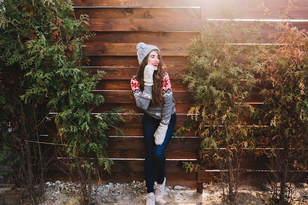 Pretty girl with long hair in knitted hat and warm gloves on wooden . She is smiling to side.