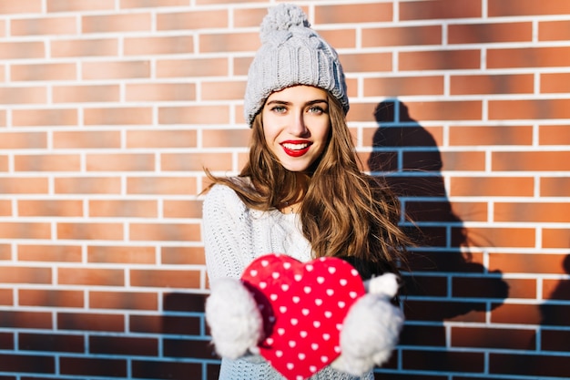 Pretty girl with long hair in knitted hat, warm sweater on wall  outside. She holds red heart in gloves, smiling .