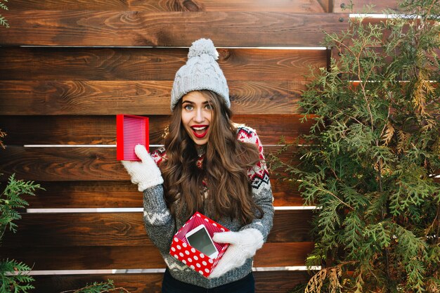 Pretty girl with long hair in knitted hat and warm sweater on wooden . She holds christmas present with phone in gloves and looks astonished .