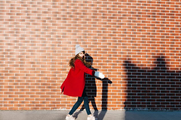 Pretty girl with long hair  in red coat and knitted hat on wall  outside. She is turning round and smiling .