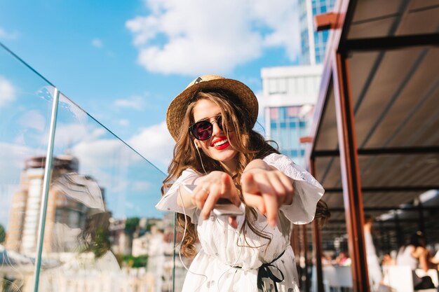 Pretty girl with long hair in sunglasses is listening to music through headphones on the terrace. She wears a white dress, red lipstick and hat . She is stretching hands to the camera. Buttom view.