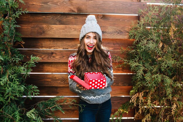 Pretty girl with long hair with christmas box on wooden . She wears warm winter clothes, knitted hat, smiling happy .