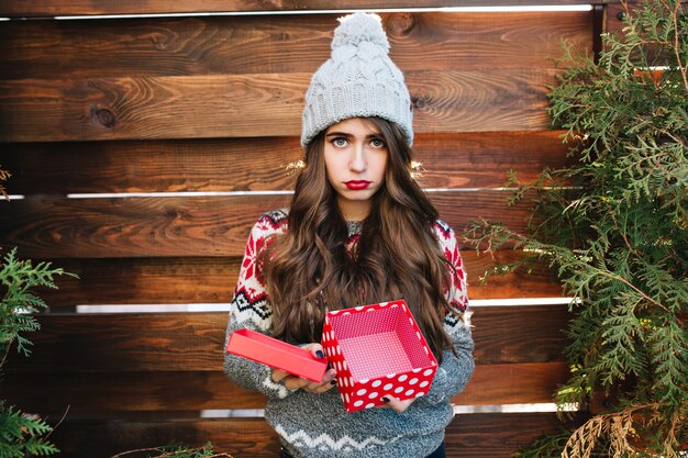 Pretty girl with long hair with empty christmas box on wooden . She wears warm winter clothes, looks dissatisfied .