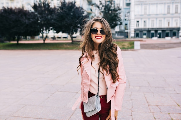 Pretty girl with long hairstyle in sunglasses is walking in city. She wears vinous pants, pink jacket, smiling .
