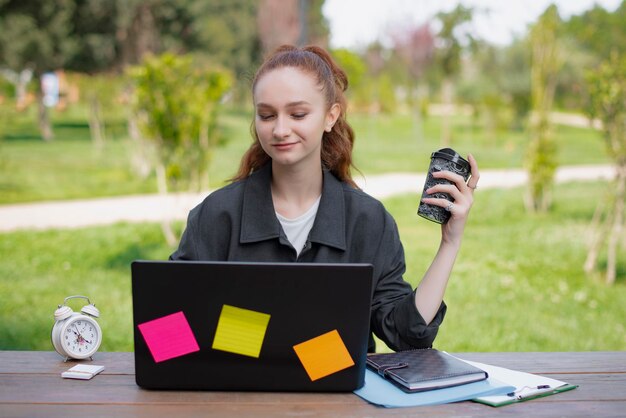 Bella ragazza con i capelli rossi che lavora alla holding del caffè del computer portatile