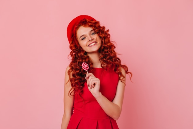 Pretty girl with wavy hair looks into camera on pink space. Woman with blue eyes dressed in red outfit posing with lollipop.