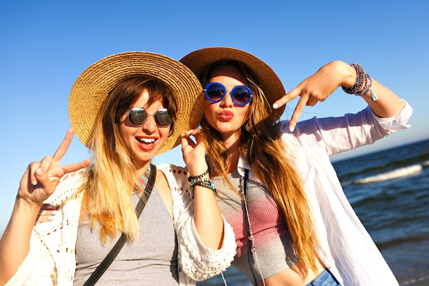 Pretty girls making selfie and sending air kisses to camera,summer traveling time, boho clothes sunglasses and straw hats.