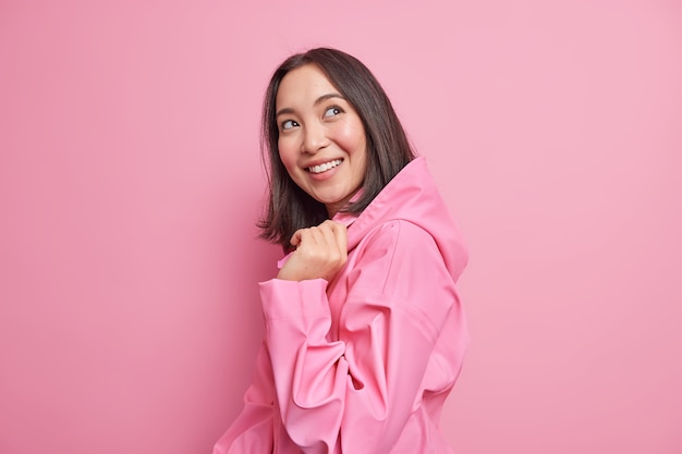 Pretty glad brunette Asian woman stands half turned against pink  wall has good mood wears stylish jacket with hood thinks about something pleasant poses happy indoor. Emotions concept