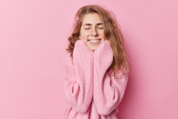 Pretty glad woman with long wavy hair closes eyes from enjoyment wears new cashmere jumper keeps hands under chin smiles gently isolated over pink background. Happy emotions and coziness concept