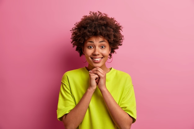 Pretty good looking girl with curly hair, keeps hands under chin, smiles gladfully, has white teeth, wears casual green t shirt, poses