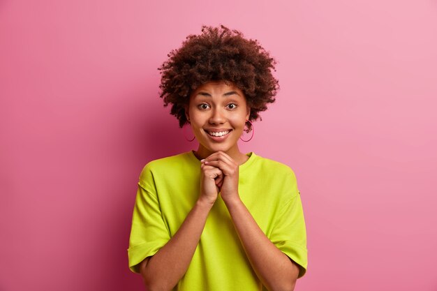 Pretty good looking girl with curly hair, keeps hands under chin, smiles gladfully, has white teeth, wears casual green t shirt, poses 