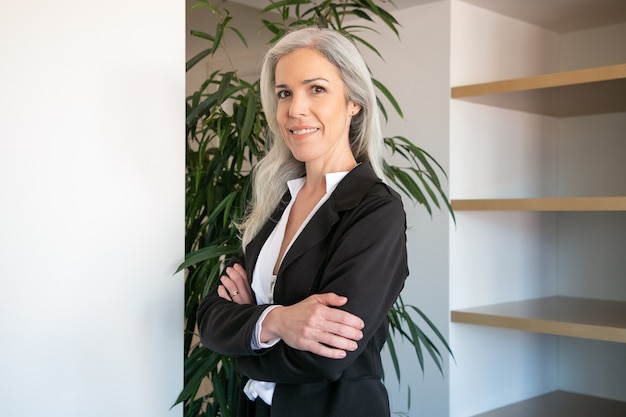 Pretty gray-haired businesswoman standing with folded hands. Portrait of confident young pretty female office employer in suit posing at work and smiling. Business, company and management concept