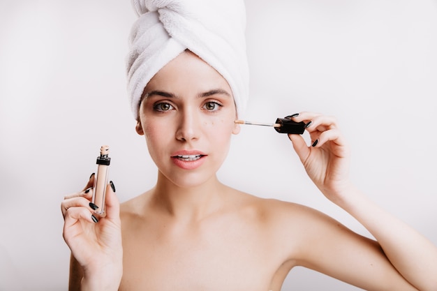 Pretty green-eyed woman on white wall. Morning routine of model that puts concealer.