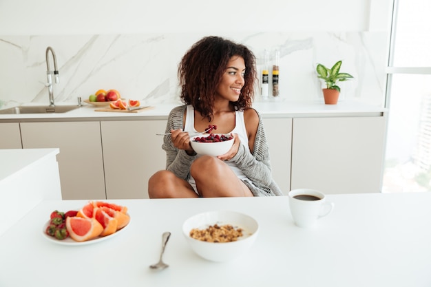 Free photo pretty happy african woman eating by the table in kitchen