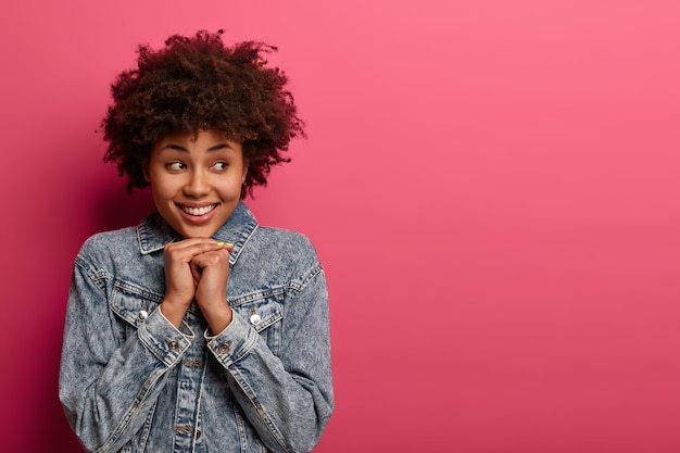 Pretty happy Afro American woman smiles positively, keeps hands under chin