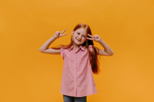 縞模様のシャツを着たかなり幸せな少女は平和のサインを示していますジーンズの陽気な子供はオレンジ色の背景に良い気分でポーズをとる