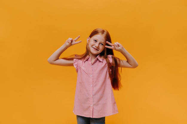 줄무늬 셔츠를 입은 꽤 행복한 소녀는 평화 기호를 보여줍니다. 쾌활한 청바지를 입은 아이는 주황색 배경에서 좋은 분위기로 포즈를 취합니다.