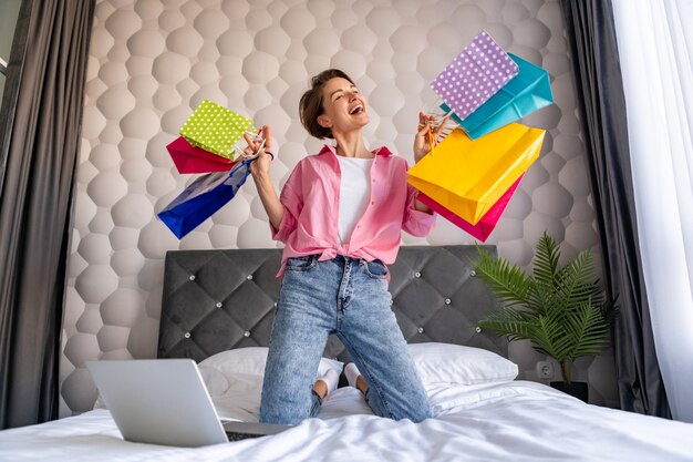 Pretty happy woman having fun jumping on bed at home with colorful shopping bags