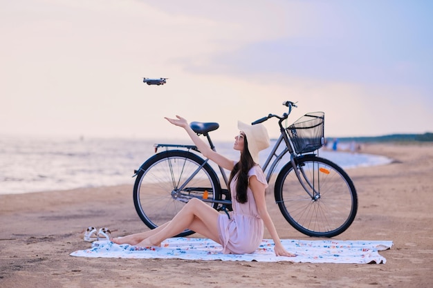 Довольно счастливая женщина пытается поймать дрон, отдыхая на пляже рядом со своим велосипедом.