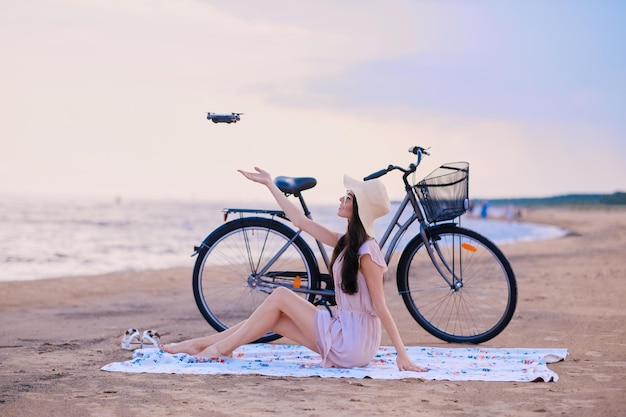 Pretty happy woman is trying to catch a drone while chilling on the beach next to her bike.