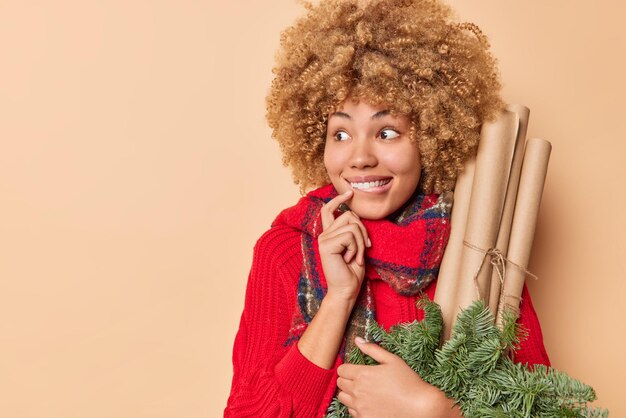 Pretty happy woman looks with happy curious expression away carries rolled paper and spruce branches for decoration smiles happily dressed in winter clothes isolated over beige wall copy space