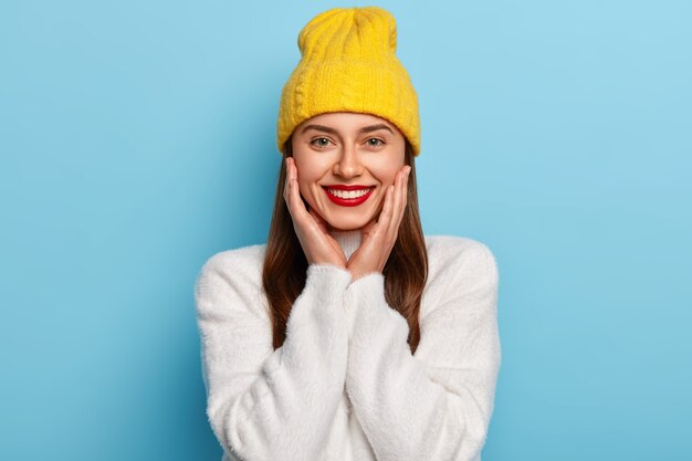 かなり幸せな女性は赤い口紅を着て、頬に手を保ち、黄色い帽子と白いカシミアのセーターを着て、青い壁にポーズをとる