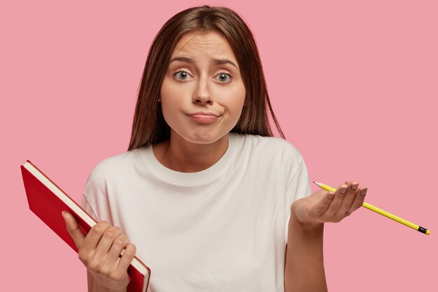 Pretty hesitant young schoolgirl has clueless doubtful expression, shruggs shoulders, holds pencil and notebook