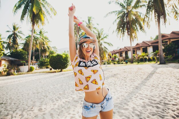 Симпатичная хипстерская женщина, идущая на пляже, танцует, слушая музыку в наушниках, в стильной красочной одежде на солнечных летних тропических каникулах в солнцезащитных очках с аксессуарами и улыбается