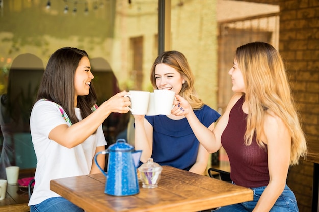 Free photo pretty hispanic female friends having fun and making a toast with coffee cups at restaurant