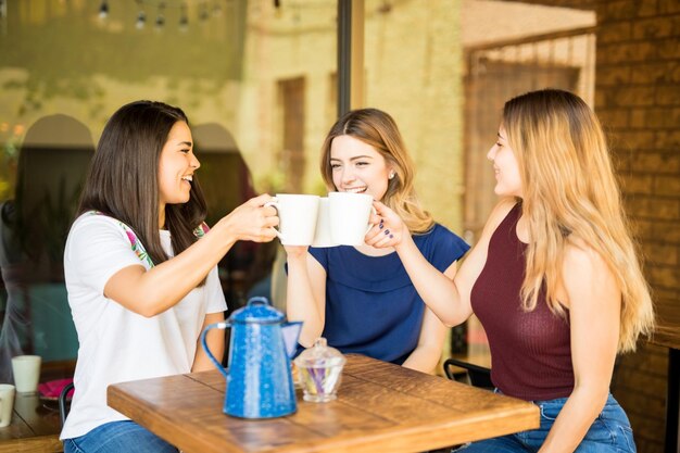 かなりヒスパニック系の女性の友人が楽しんで、レストランでコーヒーカップで乾杯をしています