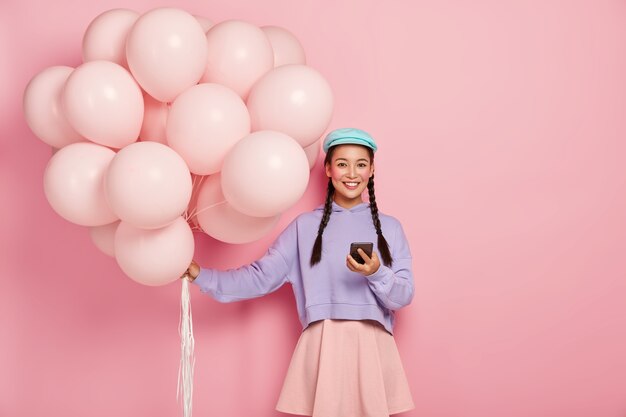 Pretty Japanese girl receives text messages on cellphone, chats online, wears stylish clothes, blue cap, stands with airballoons, being on party, smiles happily