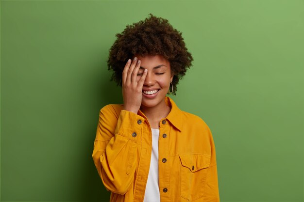 Довольно радостная темнокожая женщина держит руку на лице, смеется над смешной шуткой, закрывает глаза от радости, выражает положительные эмоции, носит модную желтую куртку, позирует в помещении над зеленой стеной.
