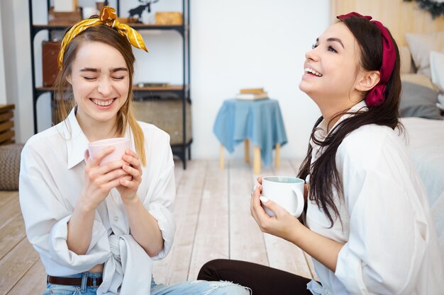 Pretty joyful girls having fun together indoors, sitting on floor with mugs of coffee
