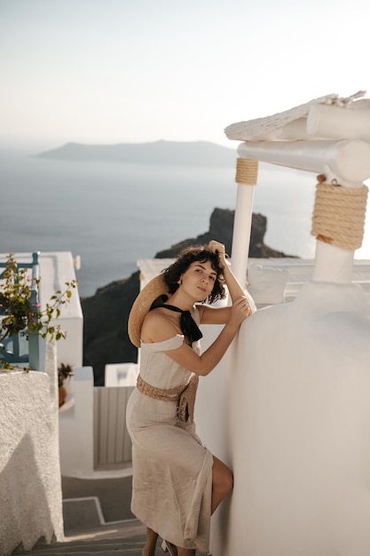 Pretty lady in beige midi dress and with straw bag leans in white wall and looks into camera outside Charming woman poses in town with sea view