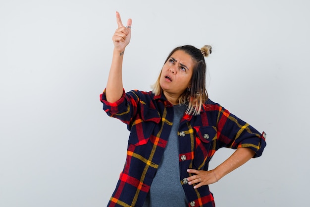 Pretty lady in casual shirt pointing up, opening mouth and looking confused , front view.