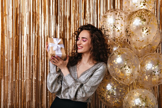 Pretty lady in good mood holding gift box on golden background