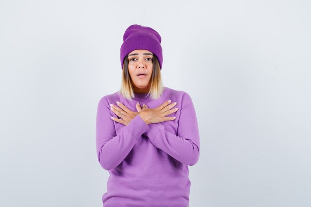 Pretty lady holding crossed hands on chest in sweater, beanie and looking puzzled , front view.