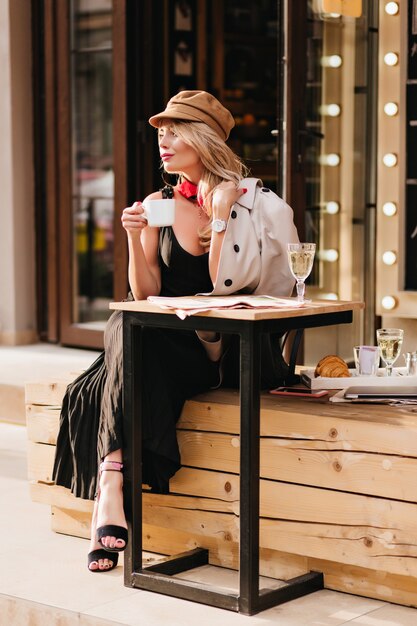 Pretty lady in long dress and black sandals enjoying lunch in outdoor cafe and looking away. Fascinating blonde girl in hat waiting friend to eat croissants together.