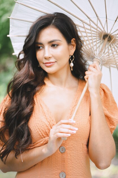 オレンジ色のドレスを着て傘を保持し、昼間は自然の中で考えているきれいな女性。