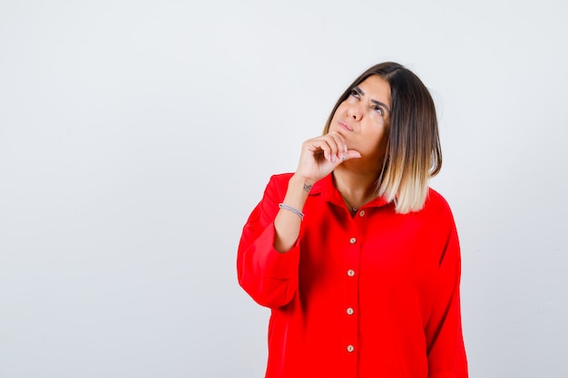 Pretty lady propping chin on hand, looking up in red blouse and looking thoughtful , front view.