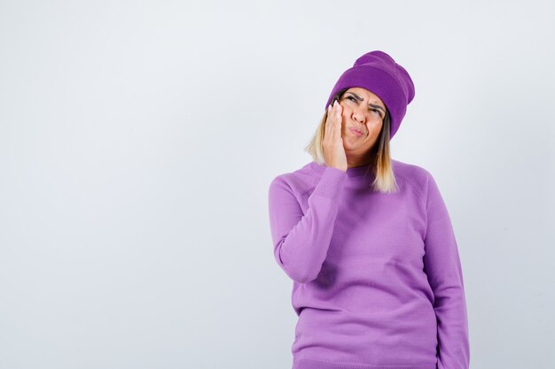 Pretty lady suffering from toothache, looking up in sweater, beanie and looking uncomfortable , front view.