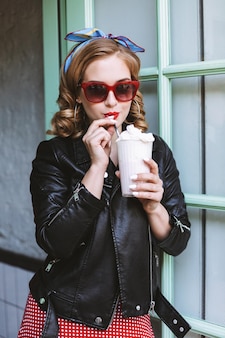 Bella signora in occhiali da sole e giacca di pelle in piedi vicino alla porta in un bar e bere milkshake