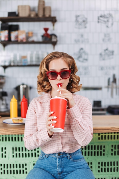 Красотка в солнцезащитных очках и рубашке сидит за барной стойкой и пьет газированную воду, проводя время в кафе