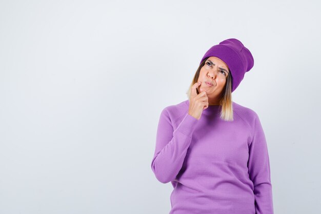 Pretty lady in sweater, beanie holding finger on cheek, looking up and looking pensive , front view.