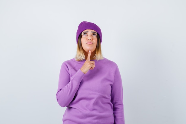 Симпатичная дама в свитере, шапочка, держащая палец под подбородком, смотрит вверх и выглядит мрачно, вид спереди.