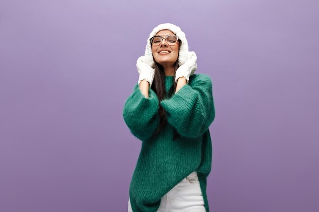 Pretty lady wearing knitted warm hat Brunette girl in white gloves eyeglasses and green sweater smiles on purple background