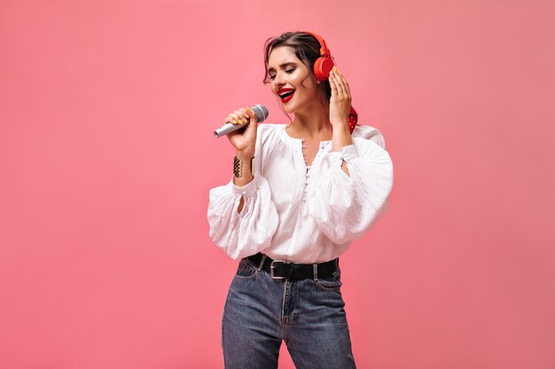 Pretty lady in white blouse sings and listens to music in headphones. Beautiful young woman in stylish outfit posing on pink background.