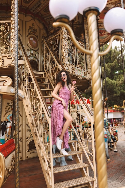 Free photo pretty lady with dark curly hair in sunglasses and dress standing with lolly pop candy in hand and happily looking aside while spending time on beautiful carousel in amusement park