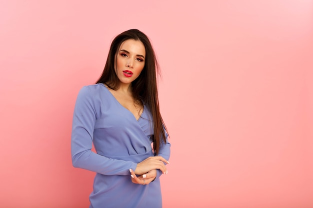 Pretty lady with red big lips and brunette long hairstyle posing on the pink background in studio. Fashionable dress on her shapely body. Decollete and long sleeves. Young sexy woman, open mouth