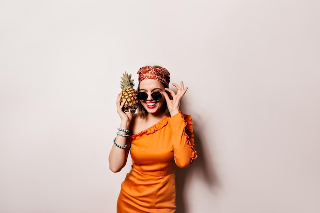 Pretty lady with red lips takes off sunglasses Woman in orange dress is smiling and holding pineapple
